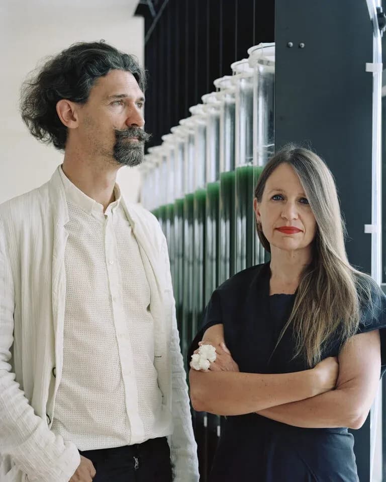A portrait of Claudia alongside co-founder Marco Poletto, with algae photobioreactors in the background—illustrating their partnership and biotech-forward architectural focus.