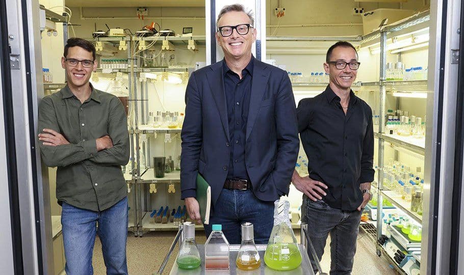 Nick standing alongside colleagues in their photobioreactor lab, with algae flasks on display—capturing their work environment